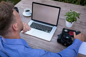Ein Mann sitzt am Holztisch mit einem Laptop, einer Kamera, Notizbuch, Kaffeetasse und einer Topfpflanze.