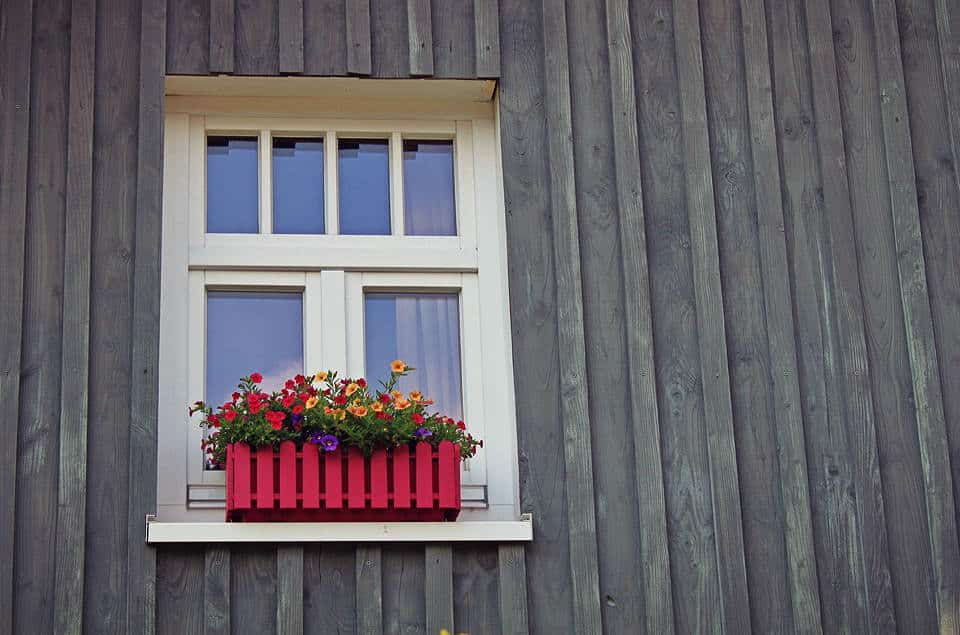 Ein weißes Fenster mit einem roten Blumenkasten an einer grauen Holzwand.