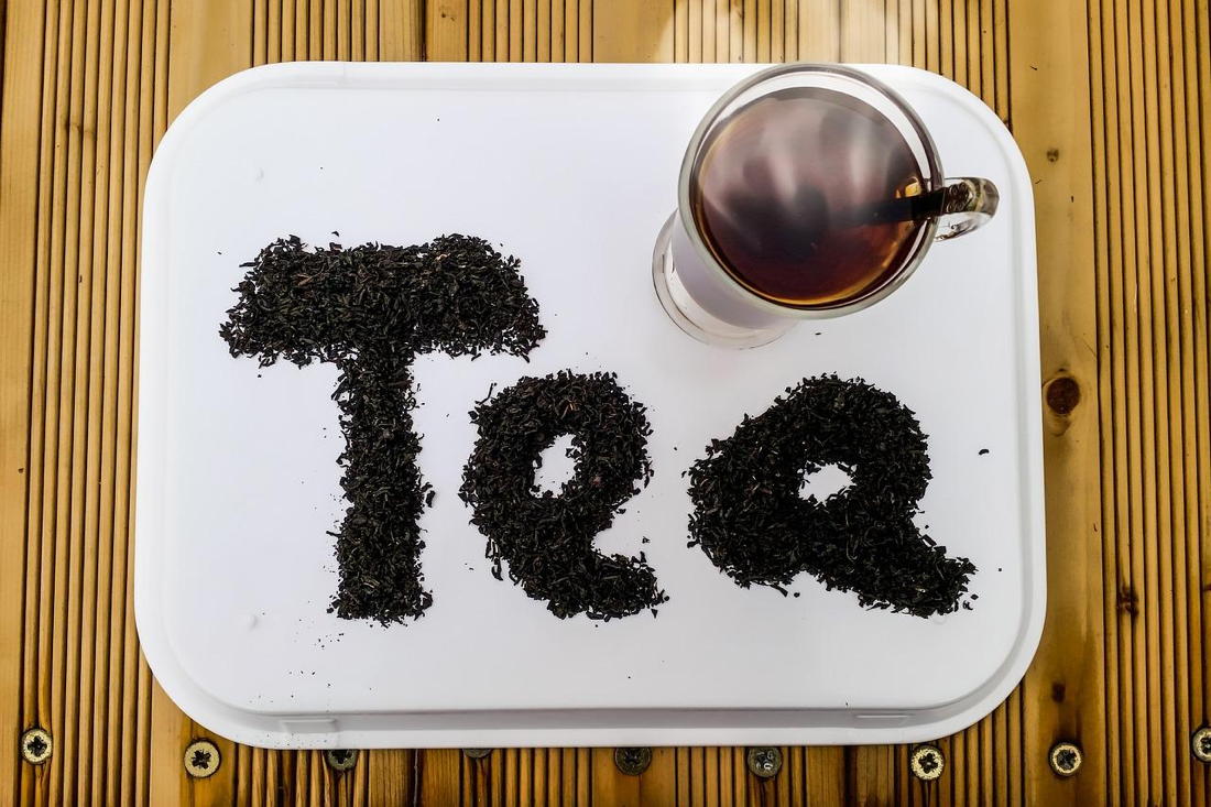 Auf dem Bild ist ein weißes Tablett mit dem Wort 'Tea', geschrieben mit Teeblättern, und daneben steht eine Tasse mit Tee.