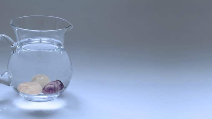 Ein Glas Krug mit klarem Wasser und darin schwebenden Steinen auf einem hellen Hintergrund.