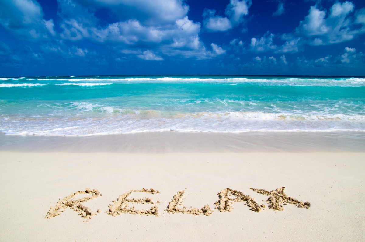 Ein tropischer Strand mit klarem blauem Wasser, weißem Sand und dem Wort 'RELAX' im Sand geschrieben.