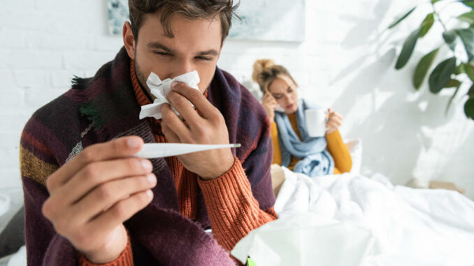 Ein Mann mit einem Fieberthermometer und einer Erkältung sitzt im Bett, während eine Frau im Hintergrund Tee trinkt.