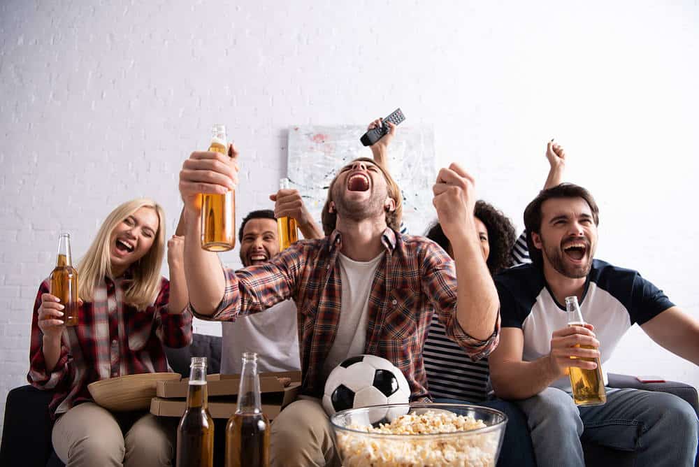 Eine Gruppe von Freunden schaut begeistert ein Fußballspiel und hält Bierflaschen in den Händen.