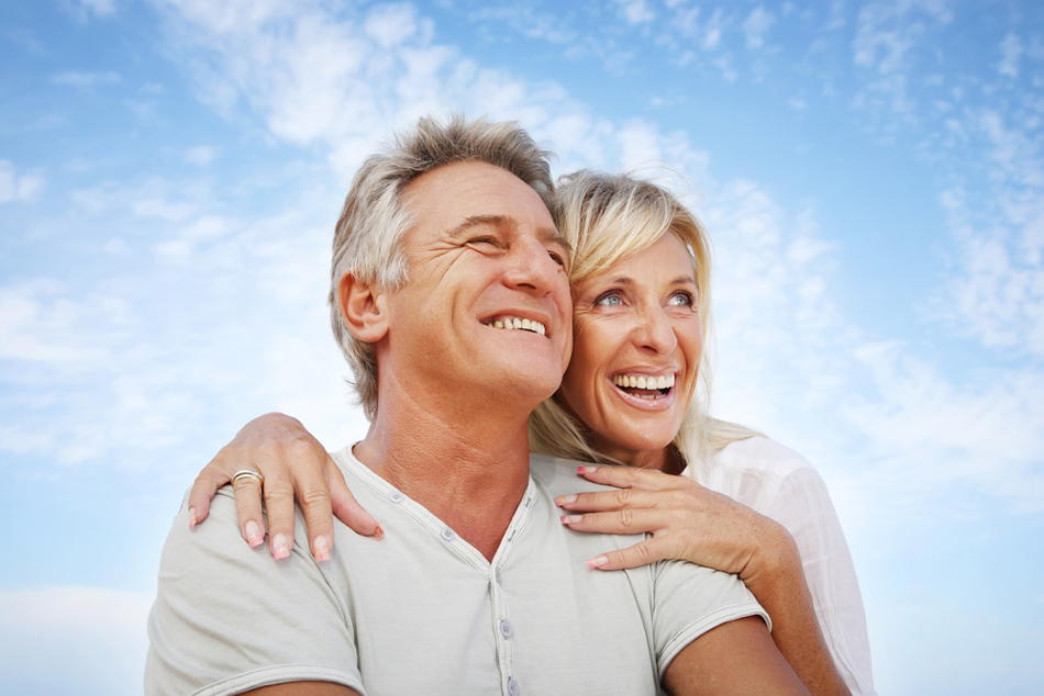 Ein älteres Paar umarmt sich lächelnd mit einem blauen Himmel im Hintergrund.