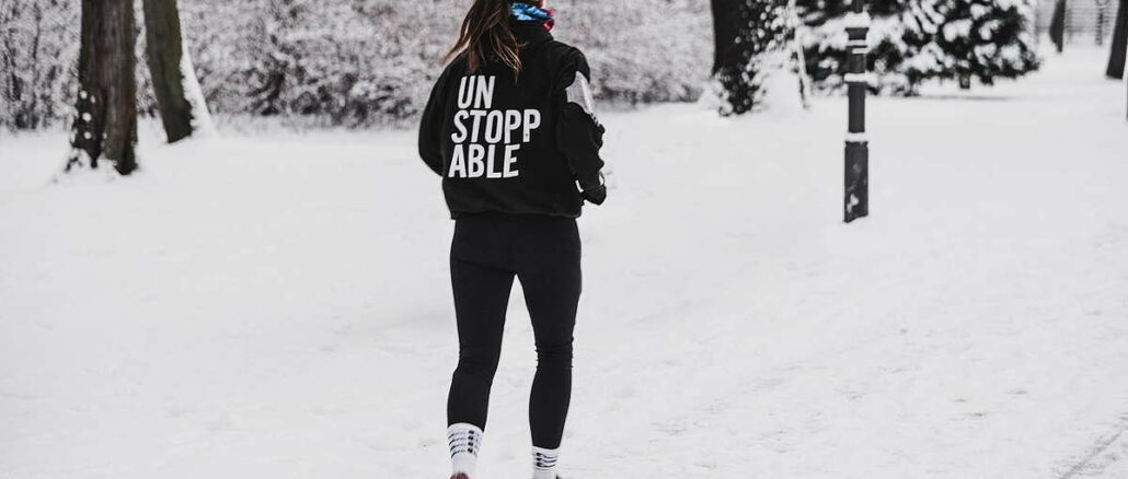 Eine Person mit einem Sweatshirt mit der Aufschrift 'UNSTOPPABLE' geht durch einen verschneiten Park.