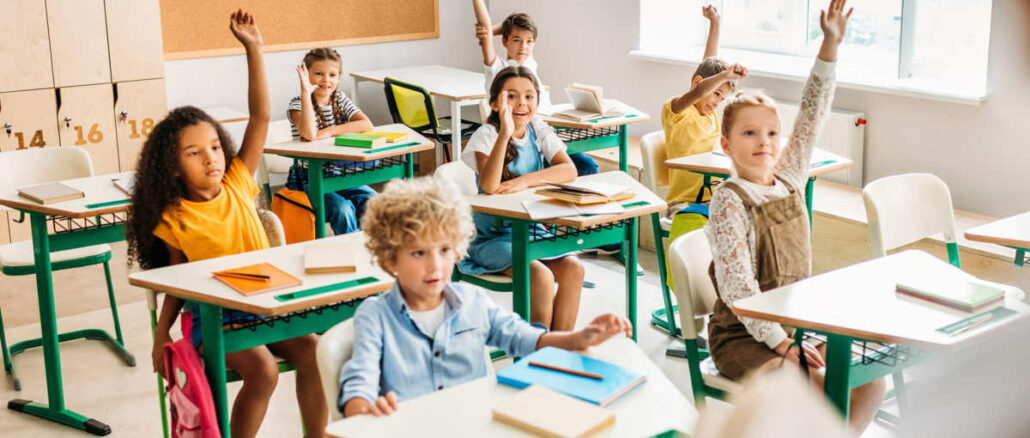 Kinder melden sich in einem hellen Klassenzimmer mit bunten Schulbänken und Heften auf den Tischen.