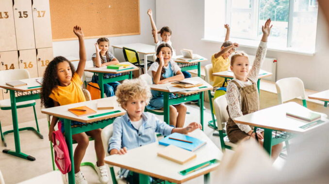 Kinder melden sich in einem hellen Klassenzimmer mit bunten Schulbänken und Heften auf den Tischen.