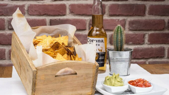 verschiedene mexikanische Speisen, Corona-Bier und kleiner Kaktus auf einem Tisch vor gemauerter Wand