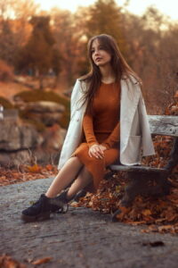 Eine Frau in einem orangefarbenen Kleid und weißem Mantel sitzt nachdenklich auf einer Bank im Herbstpark.