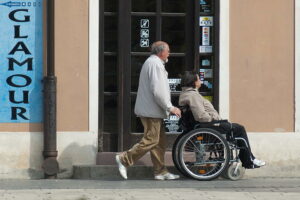 Ein Mann schiebt einen anderen im Rollstuhl an einem Geschäft mit dem Schild 'Glamour' vorbei.