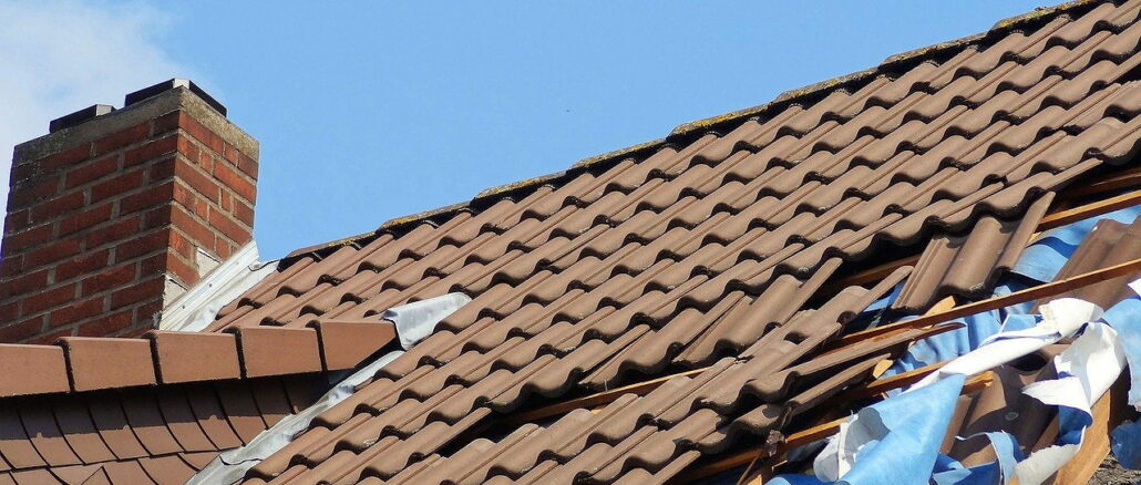 Ein beschädigtes Dach mit zerbrochenen Ziegeln und einer sichtbaren Unterlage gegen einen blauen Himmel.