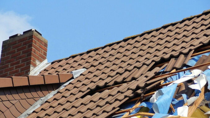 Ein beschädigtes Dach mit zerbrochenen Ziegeln und einer sichtbaren Unterlage gegen einen blauen Himmel.