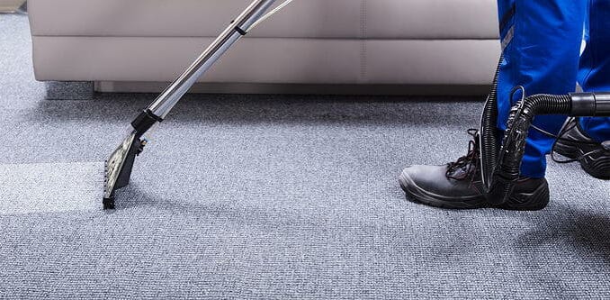 Eine Person in Arbeitskleidung reinigt mit einem professionellen Staubsauger einen grauen Teppichboden.