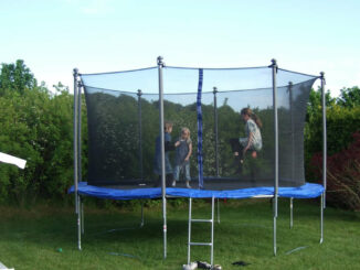 Drei Kinder spielen gemeinsam auf einem Gartentrampolin mit Sicherheitsnetz im Hinterhof.