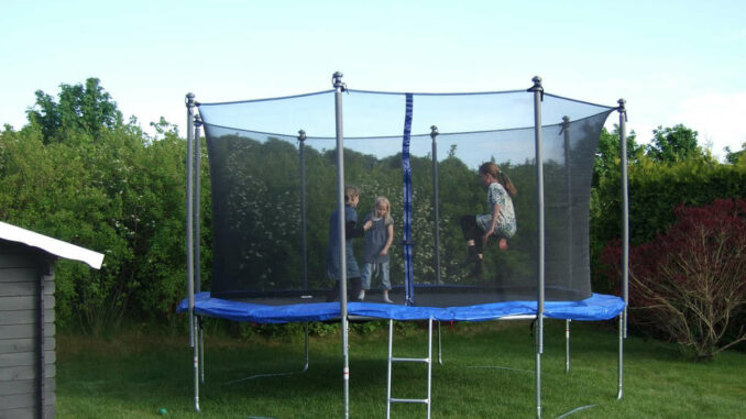 Drei Kinder spielen gemeinsam auf einem Gartentrampolin mit Sicherheitsnetz im Hinterhof.