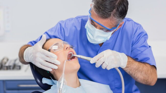 Ein Zahnarzt in Schutzkleidung untersucht die Zähne eines Patienten im Behandlungsstuhl.