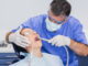 Ein Zahnarzt in Schutzkleidung untersucht die Zähne eines Patienten im Behandlungsstuhl.